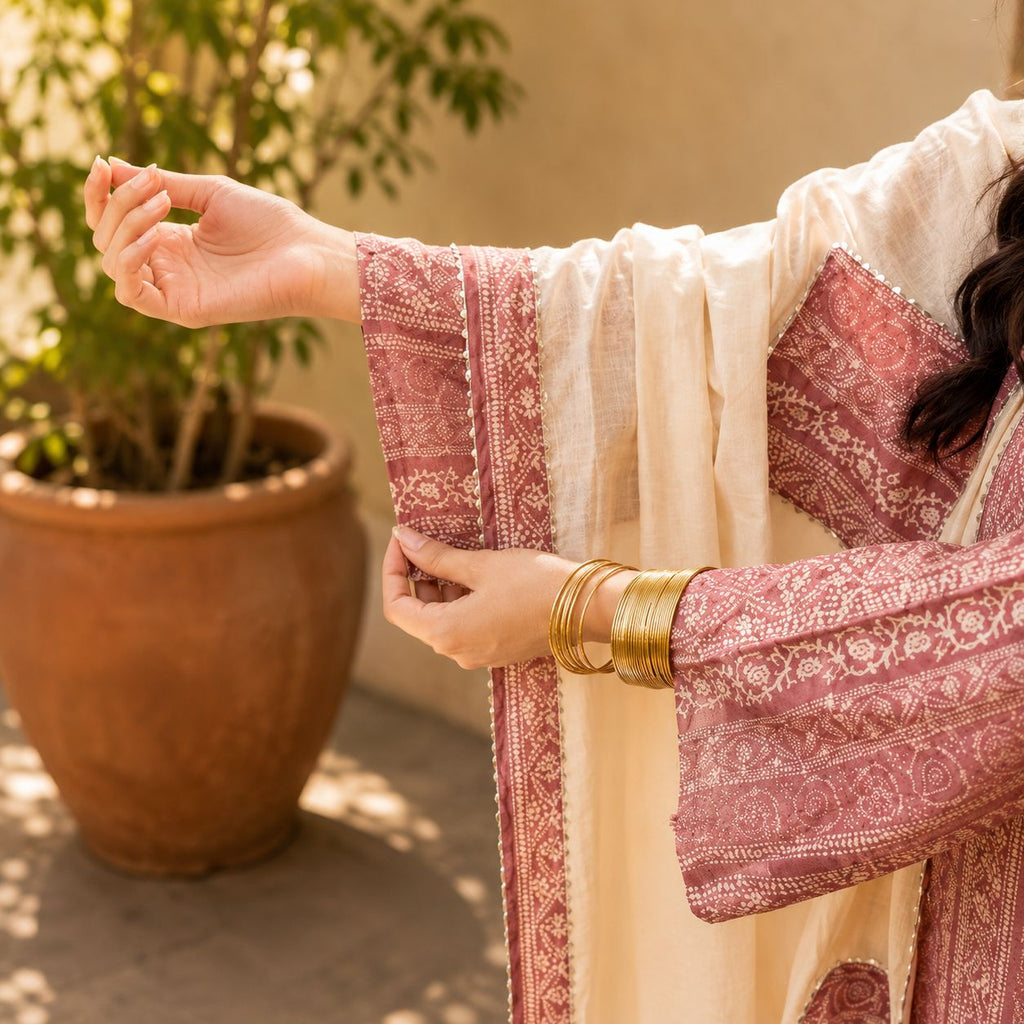 Person wearing a traditional outfit with intricate patterns, standing outdoors near a potted plant.